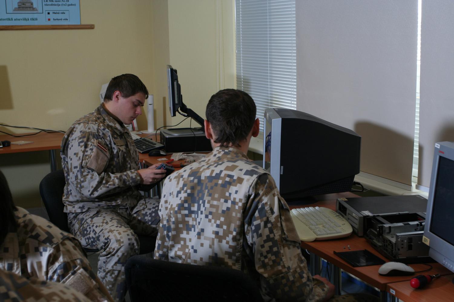 Fotogrāfijā redzami kareivis A. Maksimovs un štāba virsseržants I. Šmits lauka formā, sēžot pie galda ar datoriem un gatavojot materiālus