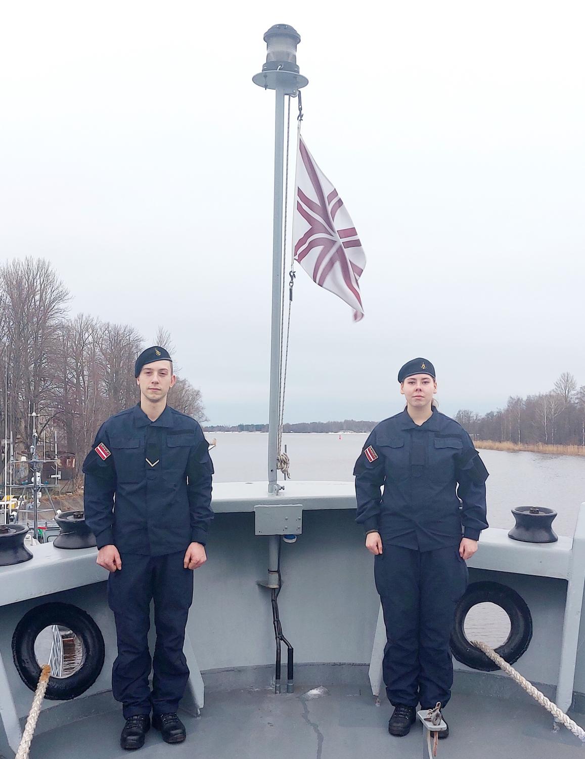Jūras spēku karavīri jaunajās zilajās formās uz kuģa un Jūras spēku karogs fonā