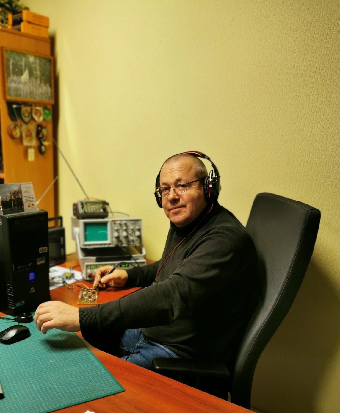 Pie datora galda sēž Jānis Stankēvičs un lodē shēmu, uz ausīm viņa ir datora austiņas
