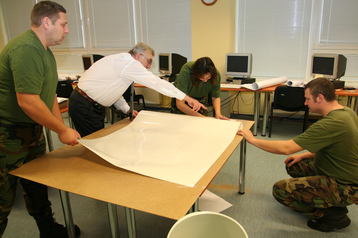 Fotogrāfijā attēloti četri karavīri lauka formās, kuri stāv pie galda ar karti un gatavo meteriālus
