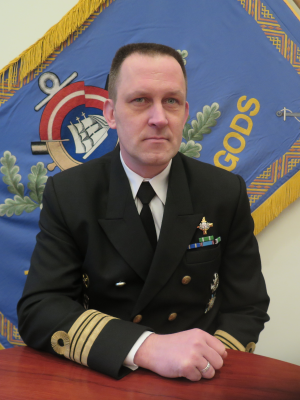 Komandkapteinis Magnuss Līcis Svētku formas tērpā uz Jūras spēku mācību centra karoga fona