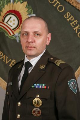 Bildē redzams štāba virsseržants Agris Priedītis. Kaujas atbalsta un nodrošinājuma mācību centra virsseržants svētku formā uz Mācību vadības pavēlniecības karoga fona.