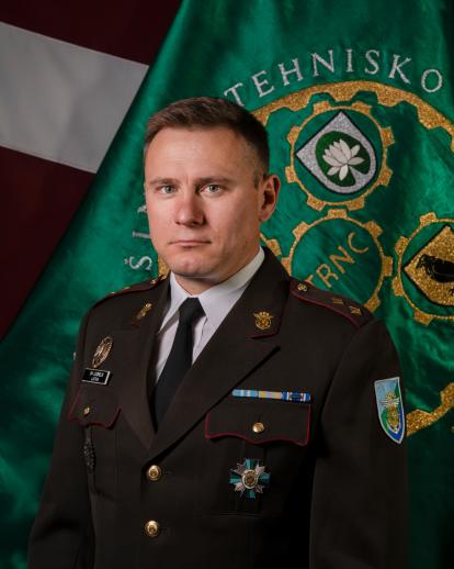 Andris Ločmelis uz vienības un Latvijas karoga fona svētku formas tērpā